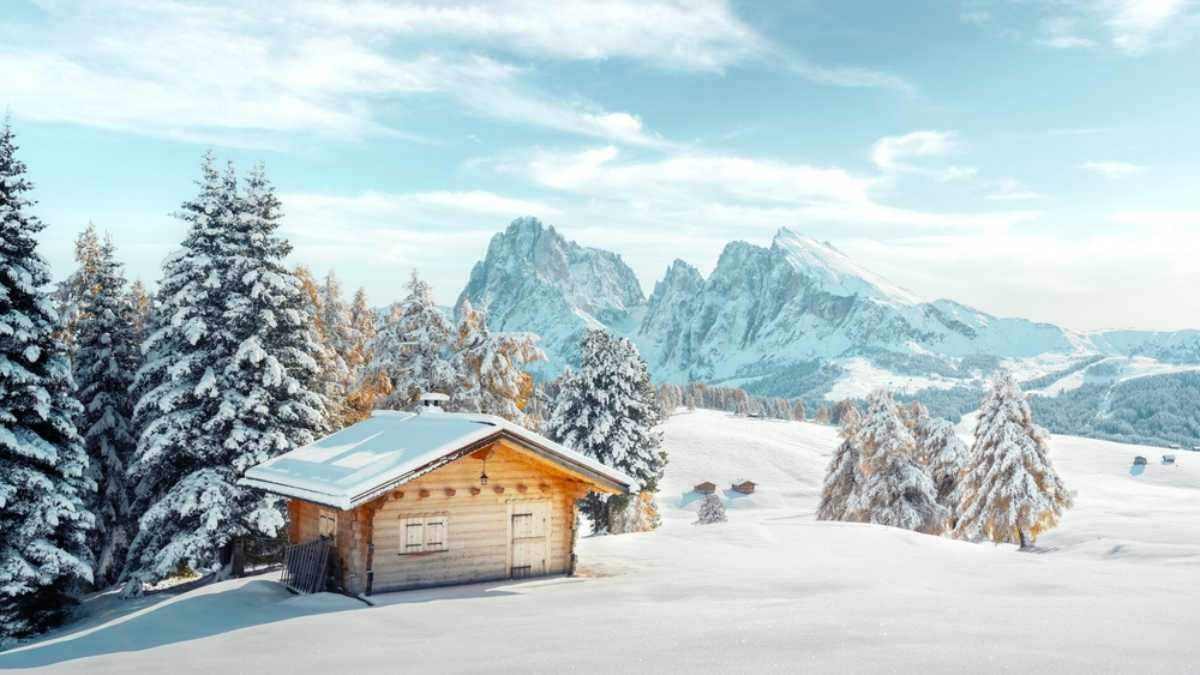 Picturesque landscape with small wooden log cabin on meadow Alpe di Siusi on winter time. Seiser Alm, Dolomites, Italy. Snowy hills with orange larch and Sassolungo and Langkofel mountains group