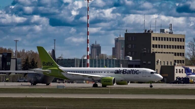 04.05.2022, Riga, Latvia. An airBaltic Airbus A220-300 taxis at Riga Airport with airport buildings and city skyscrapers in the background.