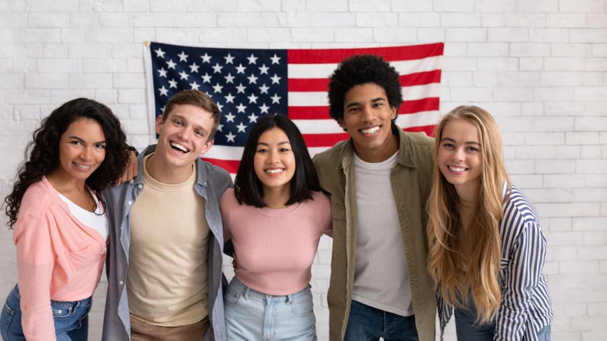 Fun education, language courses, learn english and exchange students. Young smiling people of different nationalities hug in college with large USA flag on white brick wall, free space, studio shot