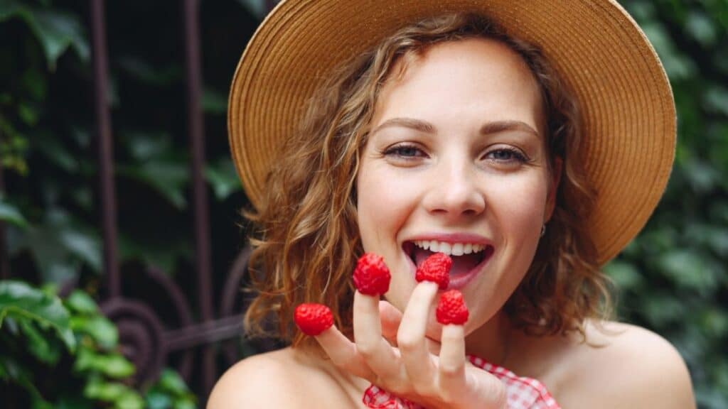 Close up young beautiful happy woman 20s wear pink dress hat put girl put raspberries on fingers eat stand outdoor near forged gate on green ivy background People urban summer time lifestyle concept.