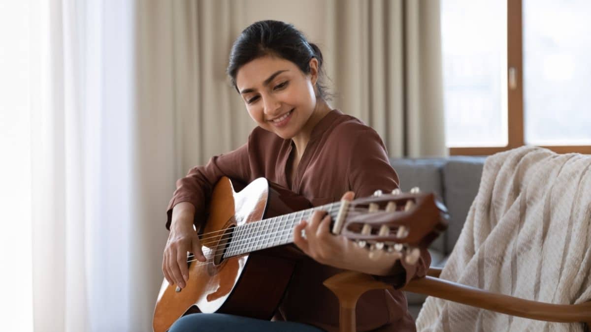 Creative hobby. Talented young mixed race female musician sit in armchair alone compose instrumental song using classic guitar. Smiling biracial lady play calm melody on musical instrument.