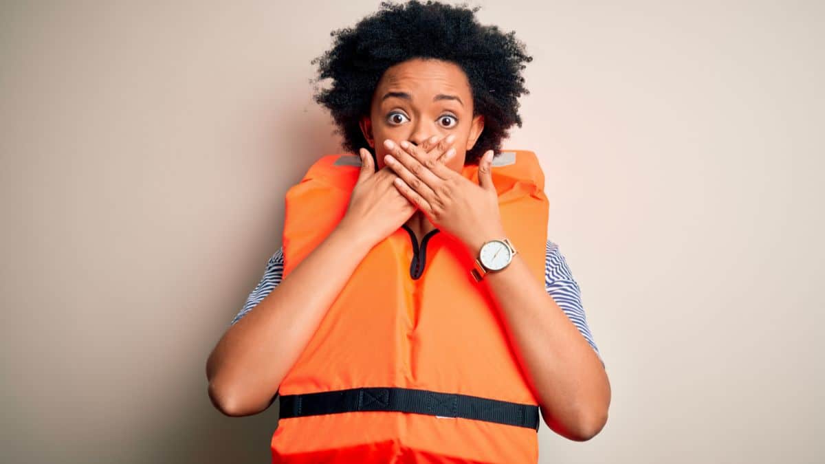 Young African American afro woman with curly hair wearing orange protection lifejacket shocked covering mouth with hands for mistake. Secret concept.