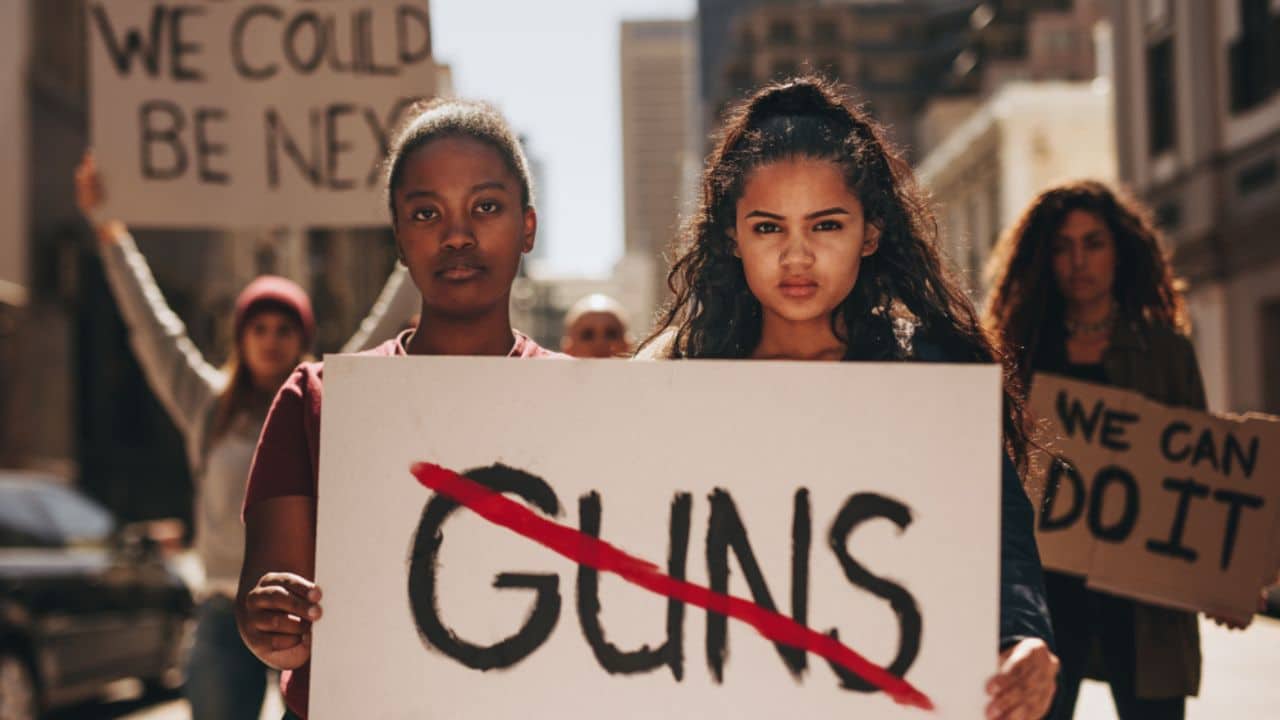 Two girls holding a banner with word guns strikethrough. Women holding sign that says not guns at a rally.