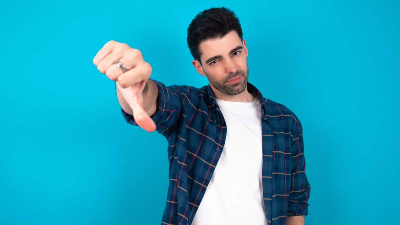 Young caucasian man wearing casual shirt over blue background feeling angry, annoyed, disappointed or displeased, showing thumbs down with a serious look