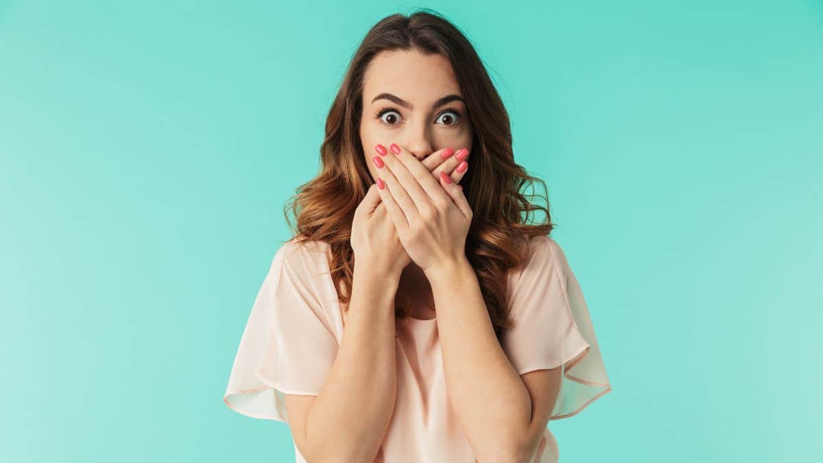 Portrait of a shocked young girl in dress looking at camera with mouth covered isolated over blue background