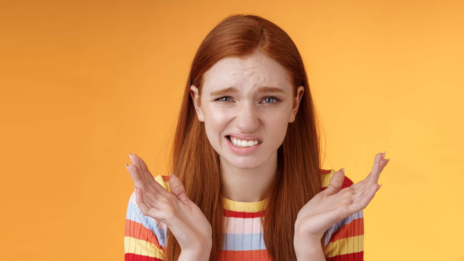 Awkward unhappy worried young redhead girl cringe feel sorry apologizing smirking smiling nervously frowning squinting spread hands sideways shrugging confused, standing orange background